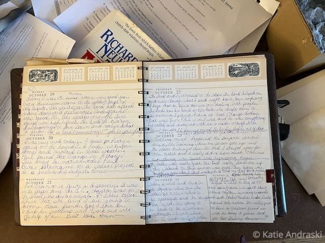 Handwritten diary entry in a spiral notebook with papers in the background.