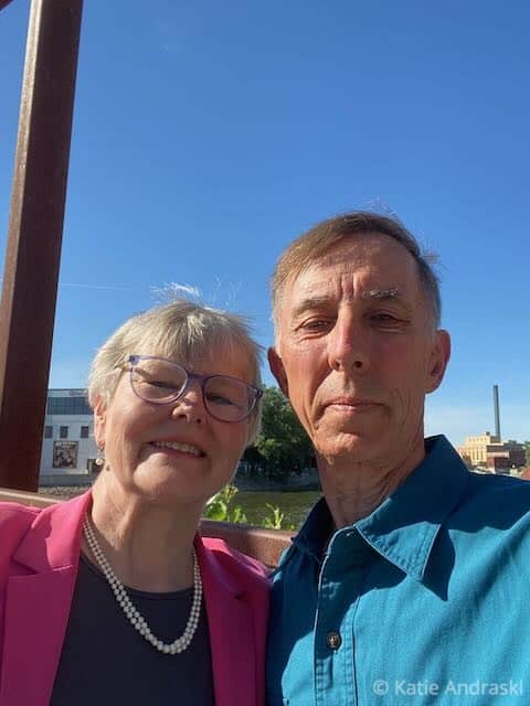An elderly couple smiling outdoors under a clear blue sky.