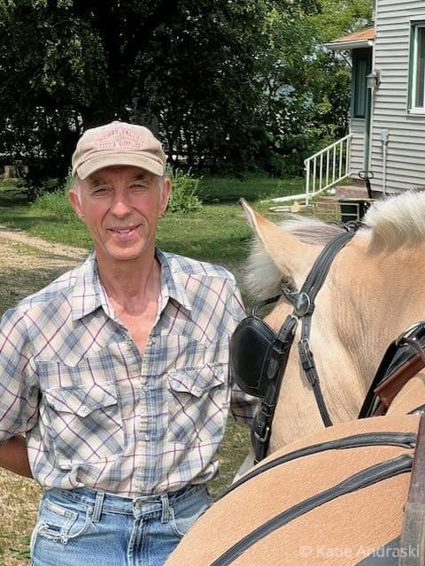 A man in a plaid shirt and cap stands beside a horse outdoors.