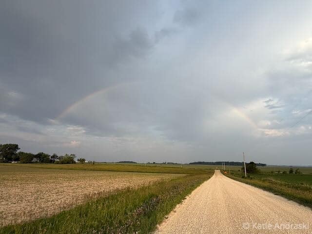A dirt road stretches through golden fields under a cloudy sky with a faint rainbow.